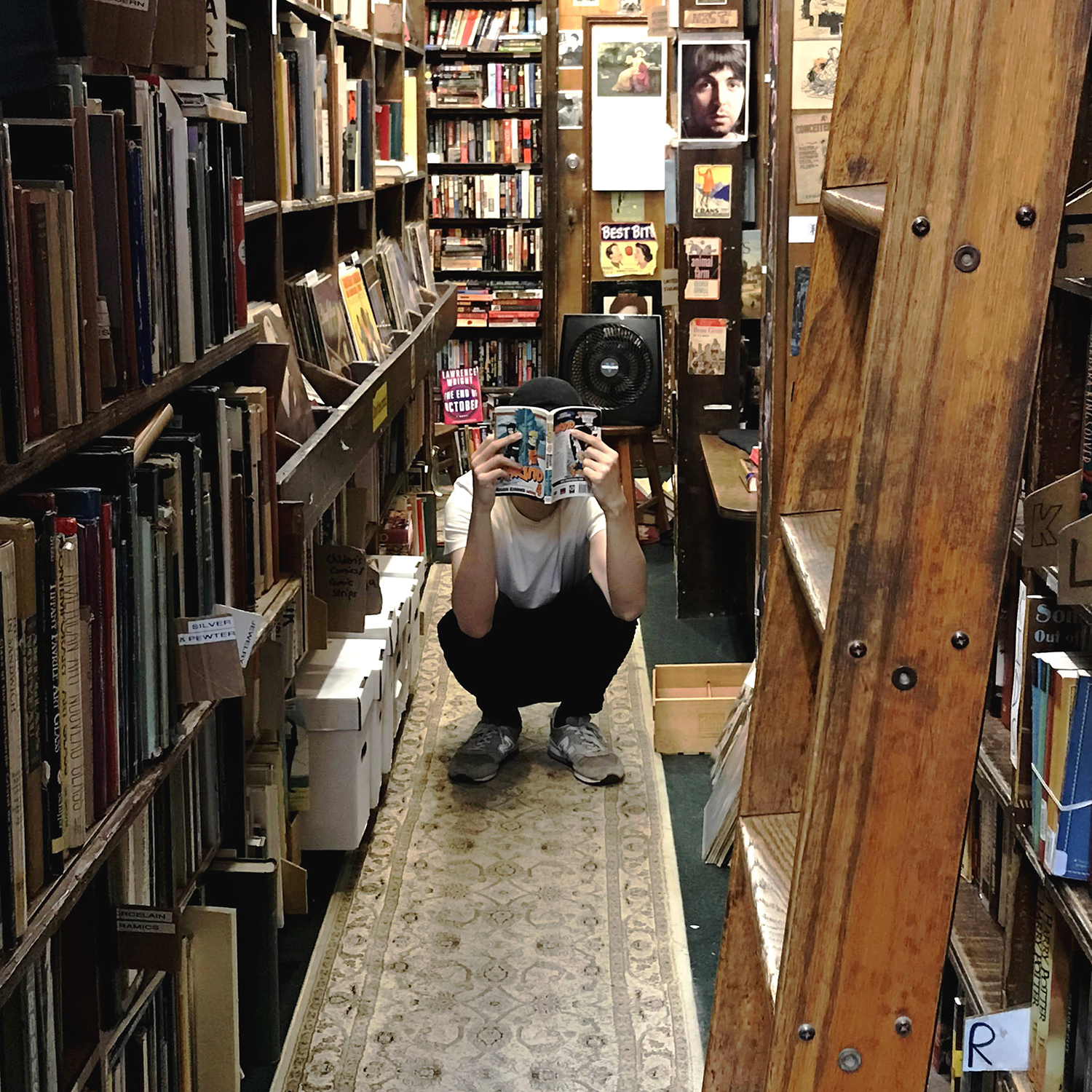 person squatting between bookshelves while holding up a book to his face
